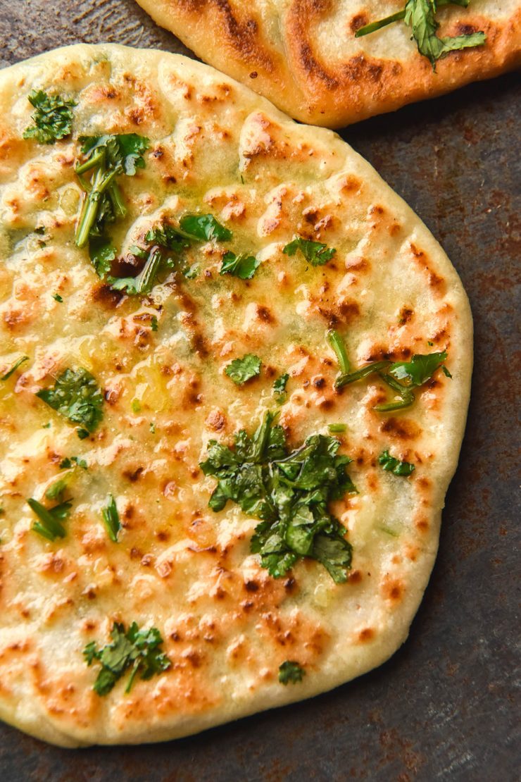 An aerial, close up view of a gluten free cheesy naan topped with garlic infused ghee, chopped coriander and sea salt flakes. A second naan sits in the top right of the image, which is set on a blue steel backdrop