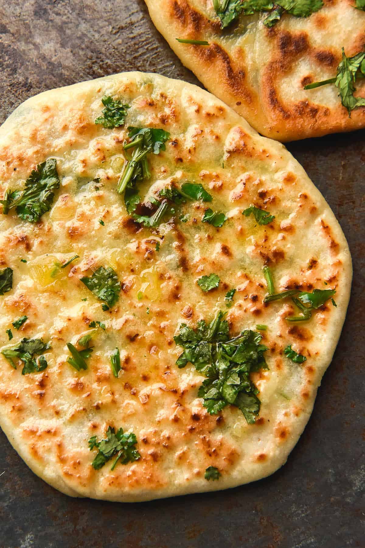 A close up image of a gluten free cheesy naan topped with garlic infused ghee, chopped coriander and sea salt flakes. The naan sits atop a dark grey backdrop and a second naan sits to the top right of the image. 