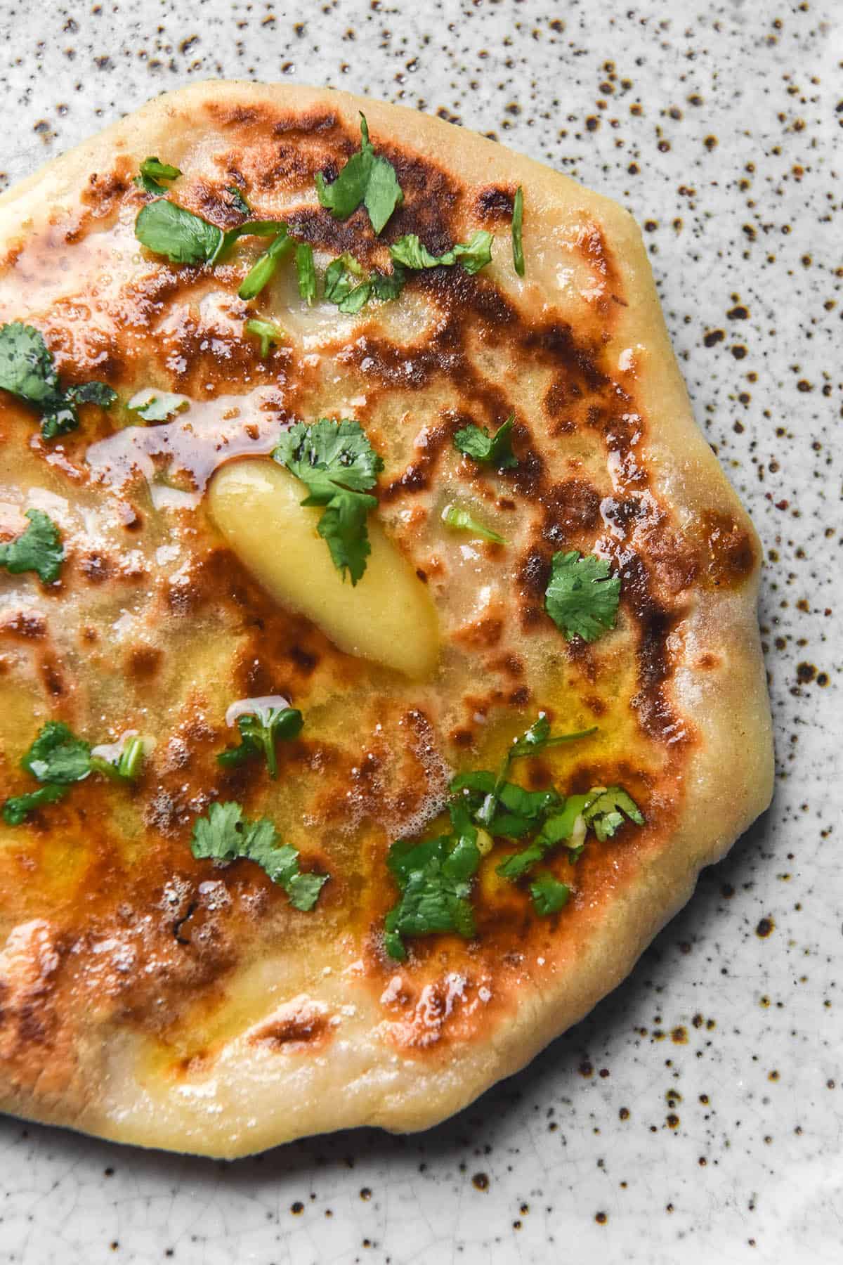 An aerial close up view of a gluten free cheesy naan atop a white speckled ceramic plate. The naan is topped with melting garlic infused ghee and chopped coriander.