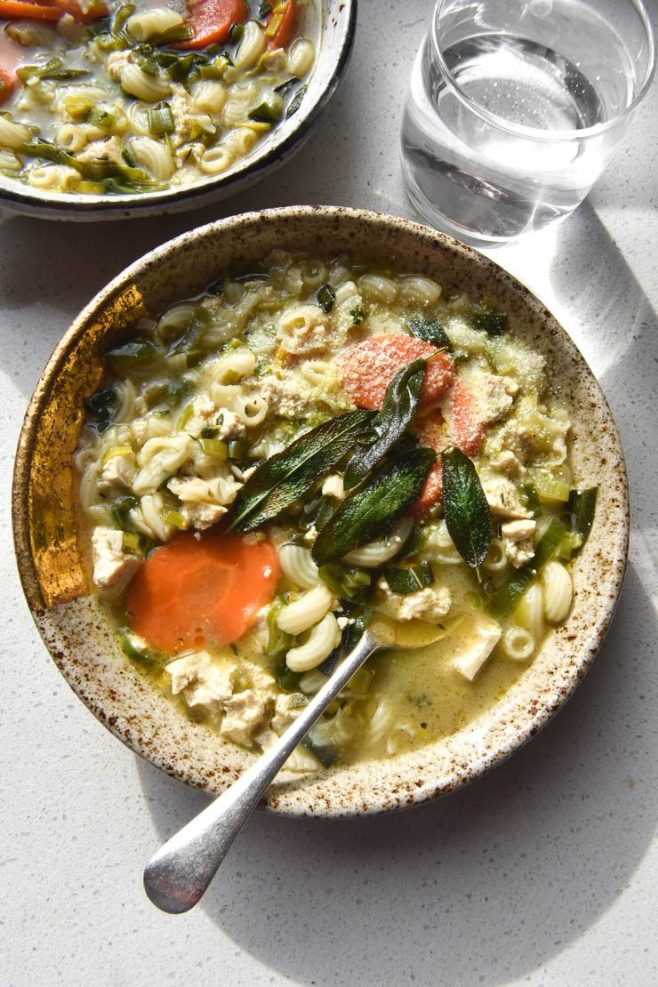 An aerial view of two speckled ceramic bowls of vegan chicken noodle soup, sitting atop a white countertop. The bowls are topped with parmesan, crispy sage leaves and truffle oil.