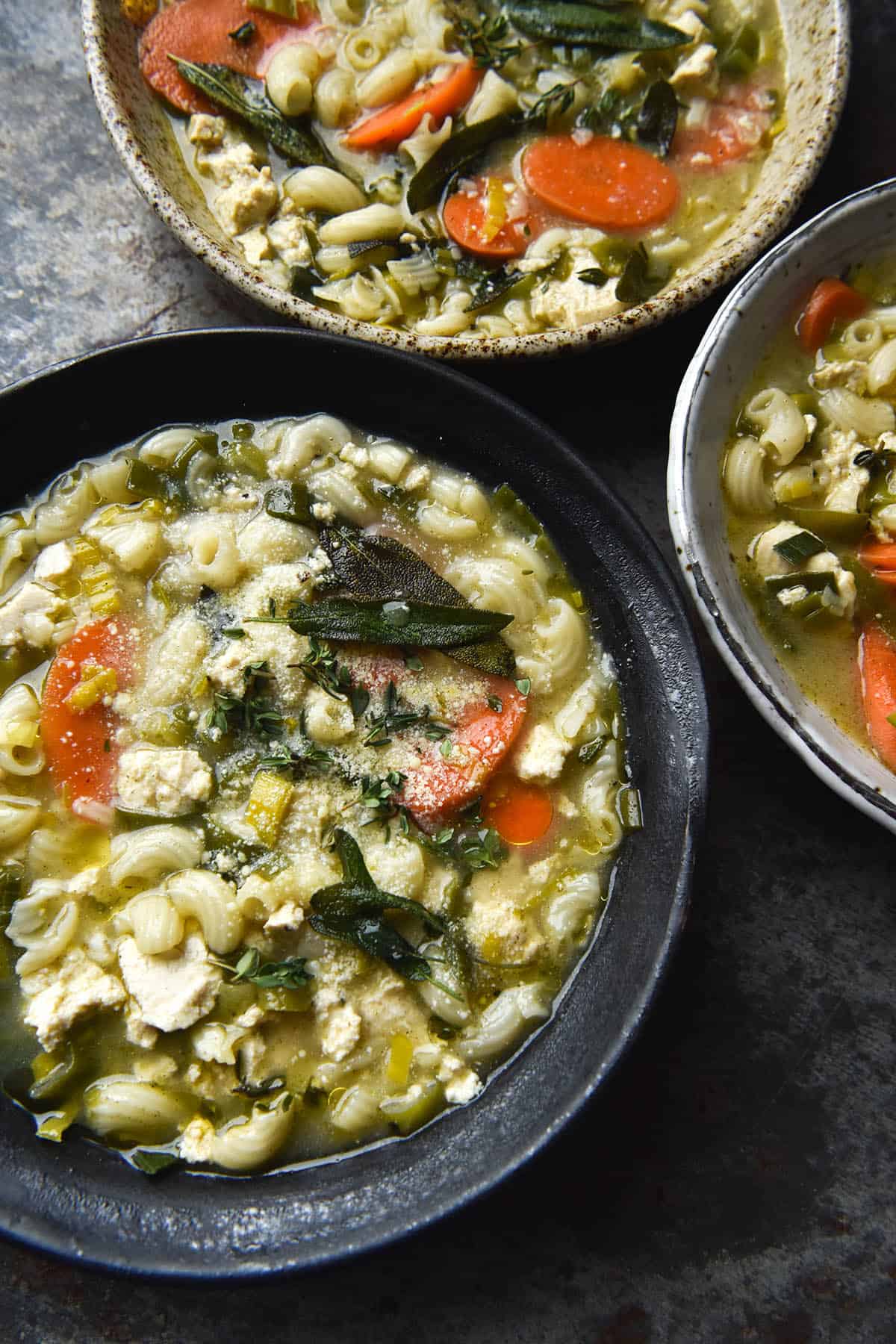 An aerial photo of three bowls of vegan chicken noodle soup atop a steel blue grey backdrop. The bowls are ceramics of various neutral toned colours and are topped with truffle oil, parmesan and crispy sage leaves. 