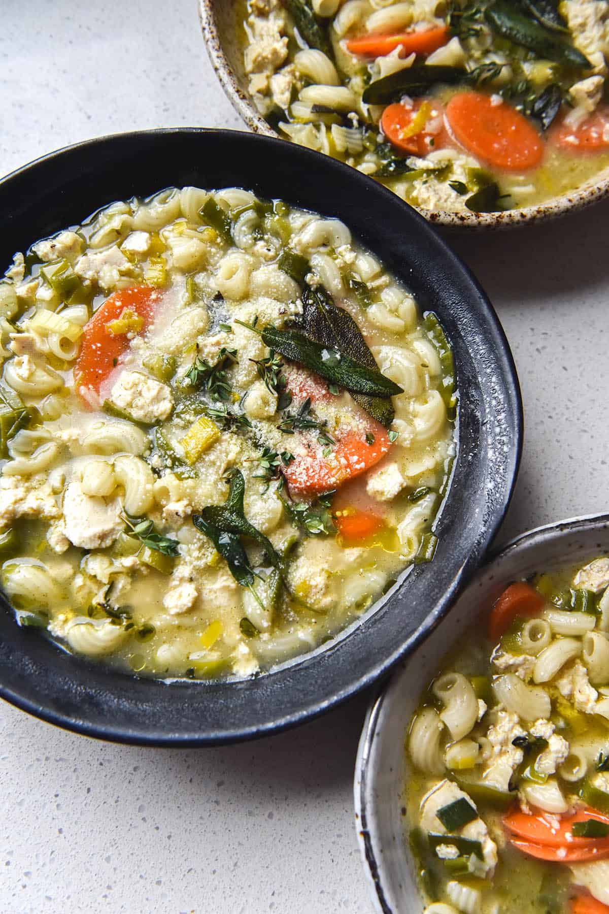 An aerial view of three bowls filled with FODMAP friendly, vegan and gluten free chicken noodle soup sitting atop a white bench top. The centre bowl is topped with parmesan, crispy sage leaves and a drizzle of truffle oil.