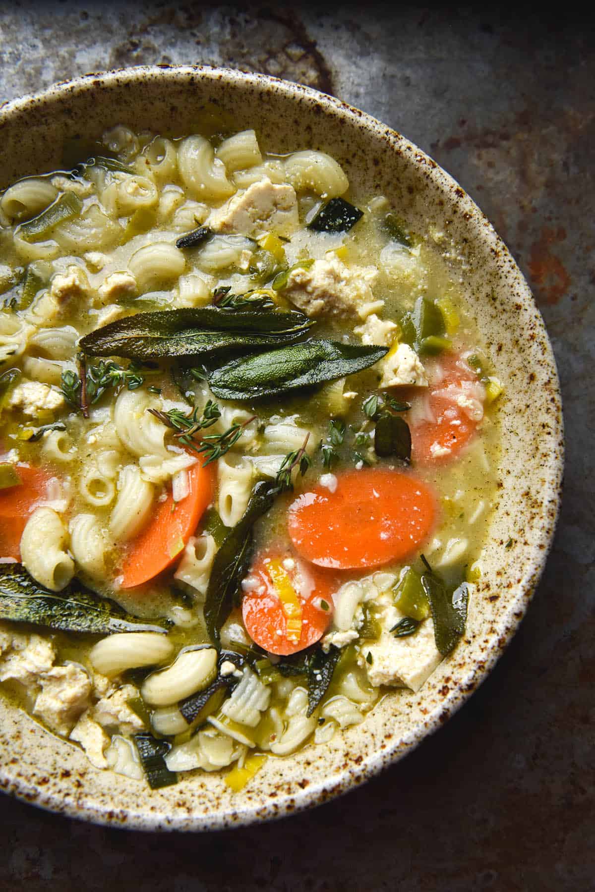An aerial close up view of a speckled beige ceramic bowl filled with vegan chicken noodle soup. Carrots, tofu cubes and pasta peek out from the broth, which is topped with crispy sage leaves, parmesan and truffle oil