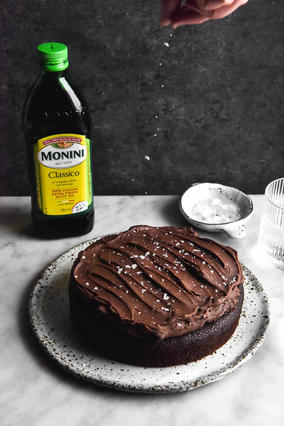 A side on image of a gluten free chocolate olive oil cake atop a white ceramic plate on a white marble table. The background is a dark blue mottled steel. A bottle of olive oil sits in the back left of the image, and a dish of sea salt flakes in the back right. A hand extends from the top of the image to sprinkle sea salt flakes down onto the cake below. 
