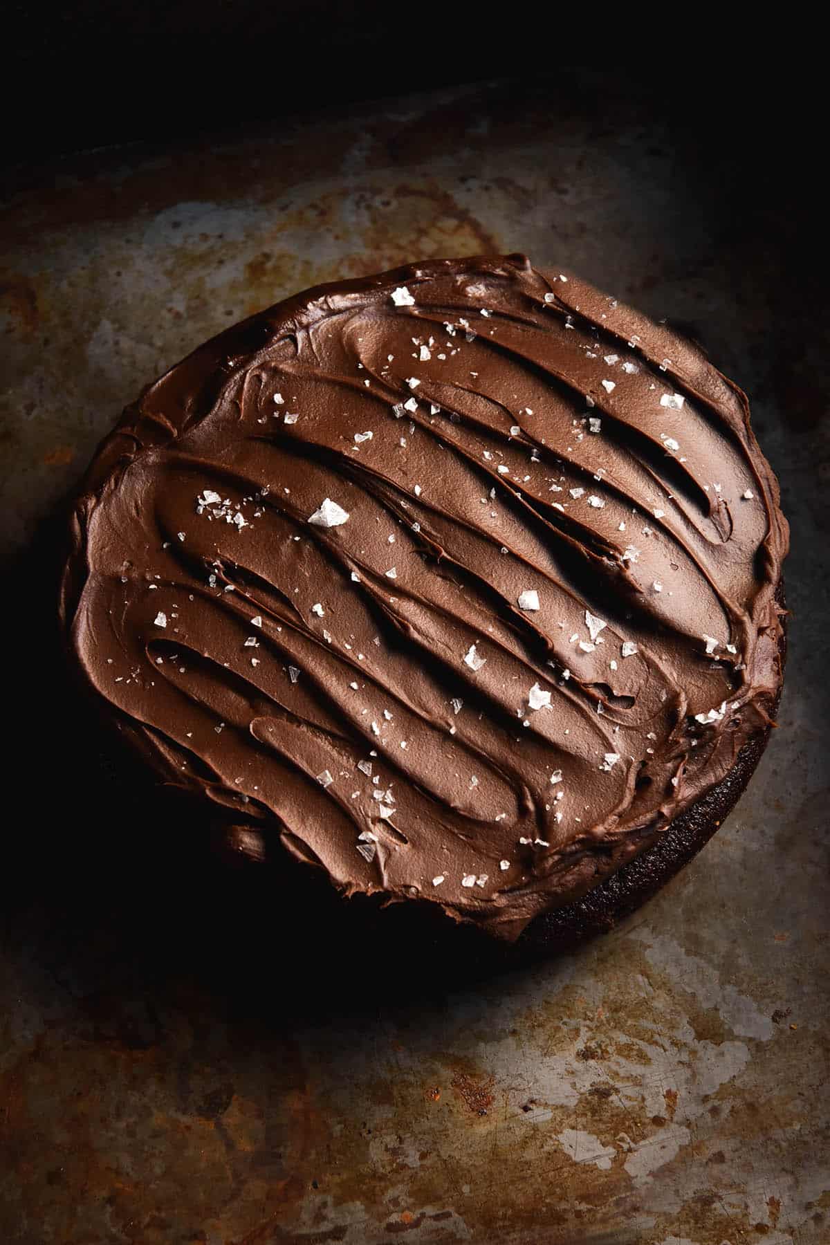 An aerial view of a gluten free olive oil chocolate cake topped with chocolate ganache and sea salt flakes. The cake sits atop a rusting dark grey backdrop.
