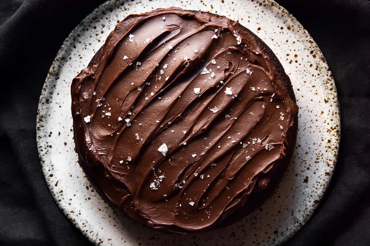 An aerial view of a gluten free olive oil chocolate cake topped with chocolate ganache and sea salt flakes. The cake sits atop a white speckled ceramic plate which sits on a black linen tea towel.