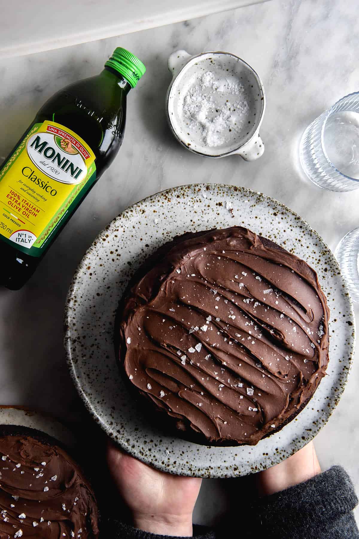 An aerial view of a gluten free chocolate olive oil cake topped with chocolate ganache and sea salt flakes. The cake sits atop a white speckled ceramic plate on a white marble table, surrounding by water glasses and an olive oil bottle. Two hands hold up the ceramic plate from the bottom right of the image.