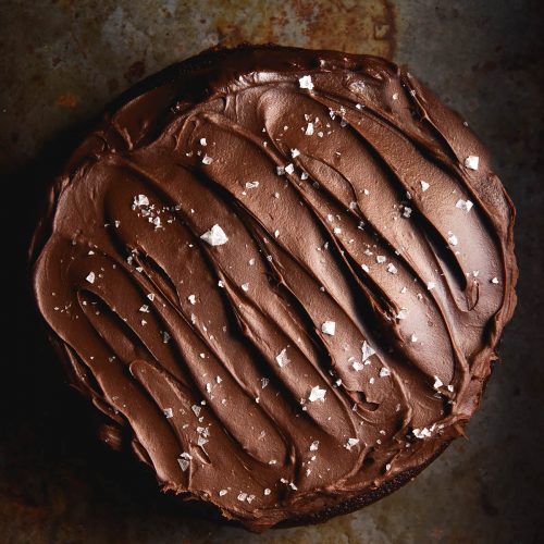 An aerial image of a gluten free chocolate cake topped with chocolate ganache and sea salt flakes on a dark steel backdrop