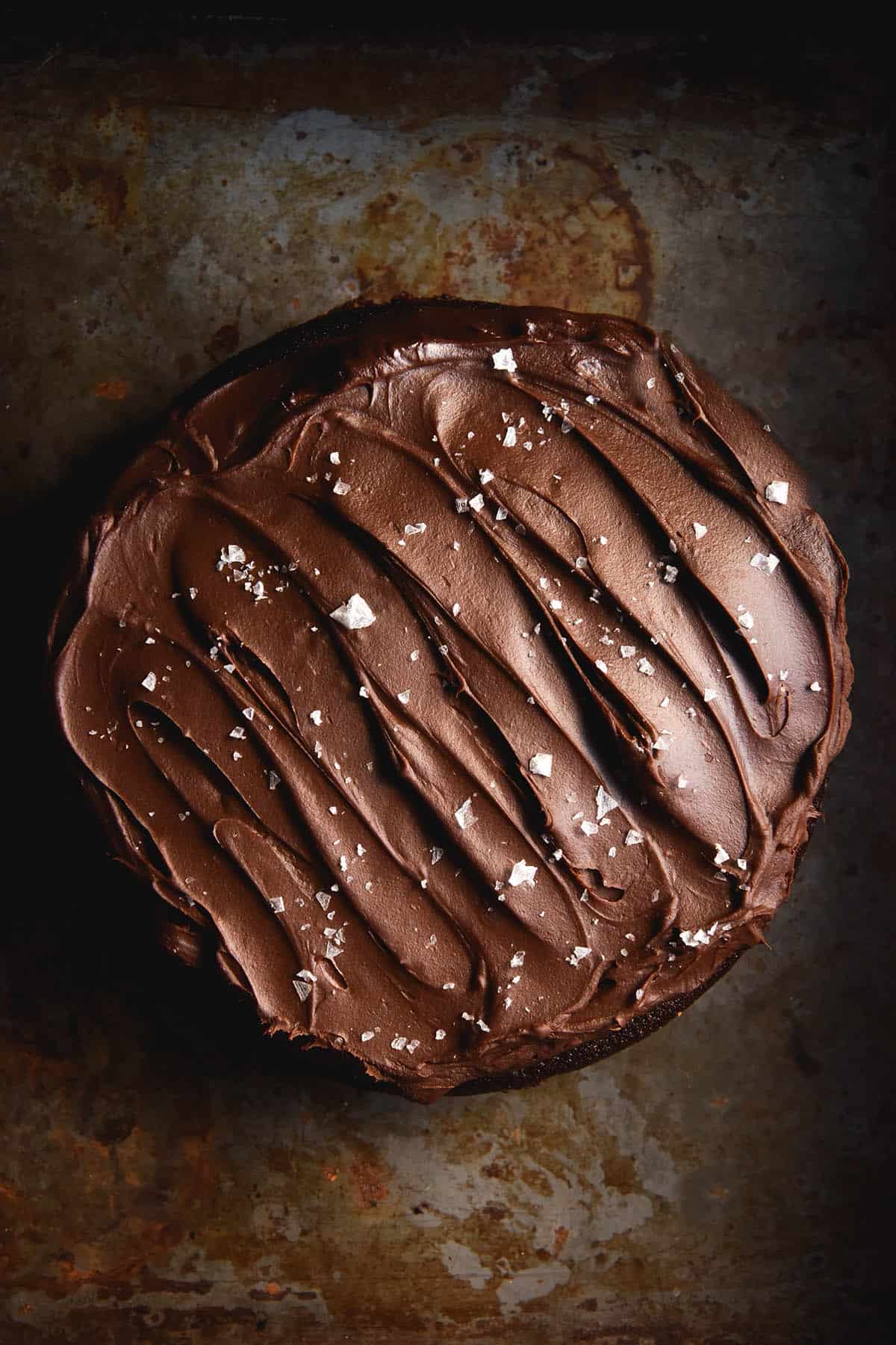 An aerial view of a gluten free olive oil chocolate cake topped with chocolate ganache and sea salt flakes. The cake sits atop a rusting dark grey backdrop.