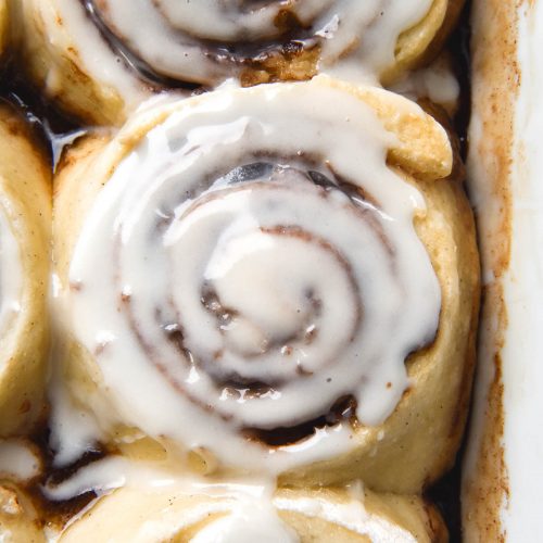 An aerial view of gluten free, vegan cinnamon scrolls sitting snug in a baking dish. The scrolls have been topped with a glaze that oozes into the cinnamon swirls