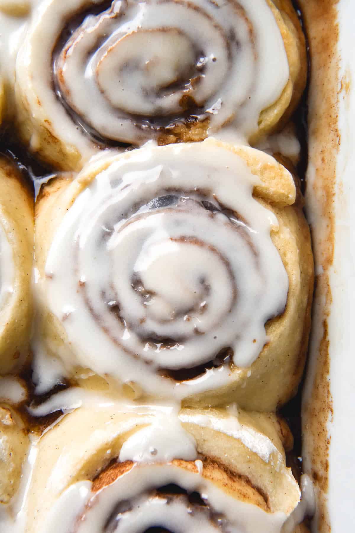 An aerial view of gluten free, vegan cinnamon scrolls sitting snug in a baking dish. The scrolls have been topped with a glaze that oozes into the cinnamon swirls