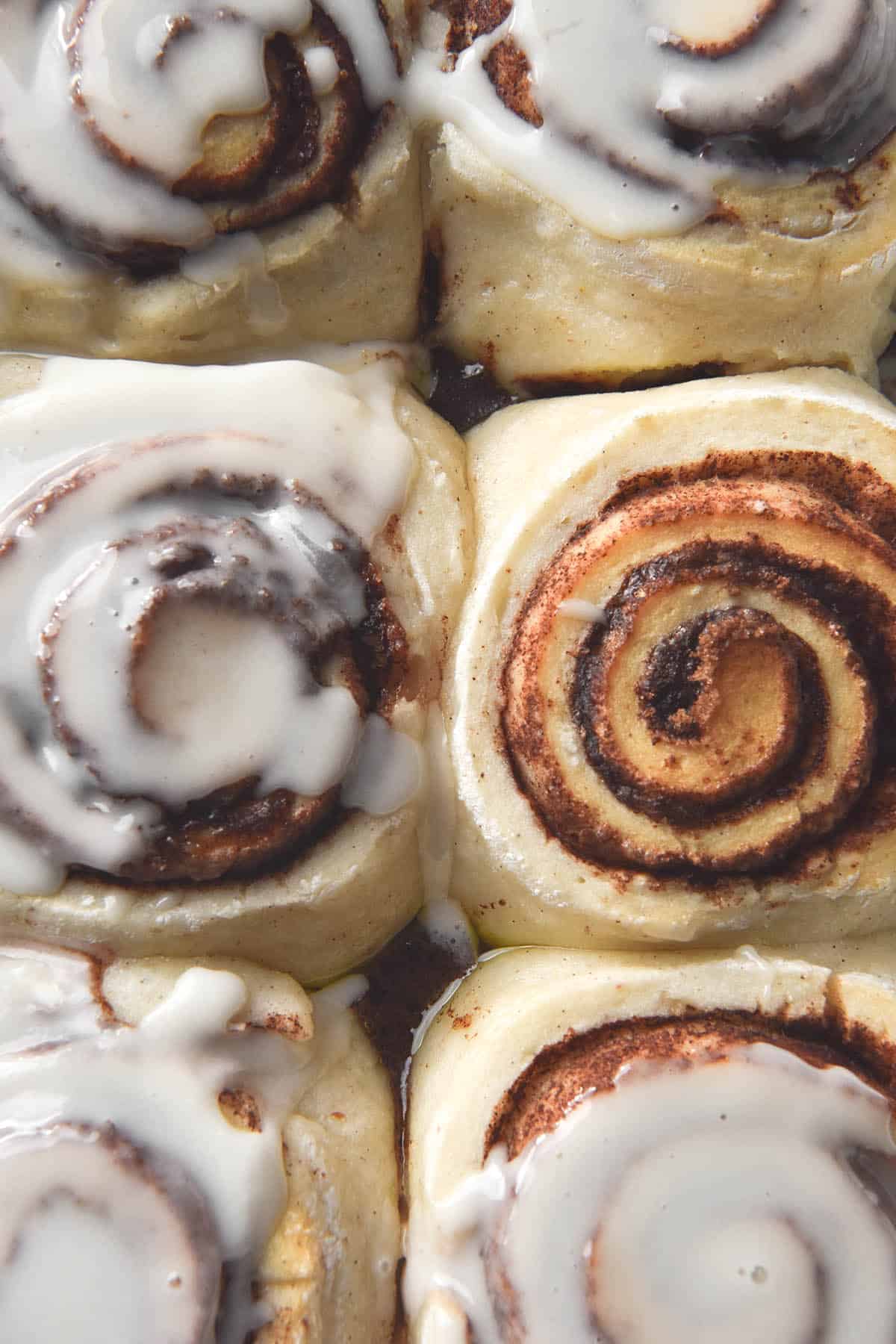 An aerial view of a tray of gluten free, vegan and yeast free cinnamon scrolls. Six scrolls sit snug in the tray with rich swirls of cinnamon and brown sugar. Most of the scrolls have been drizzled with a cinnamon scroll glaze, which melts into the scroll. 