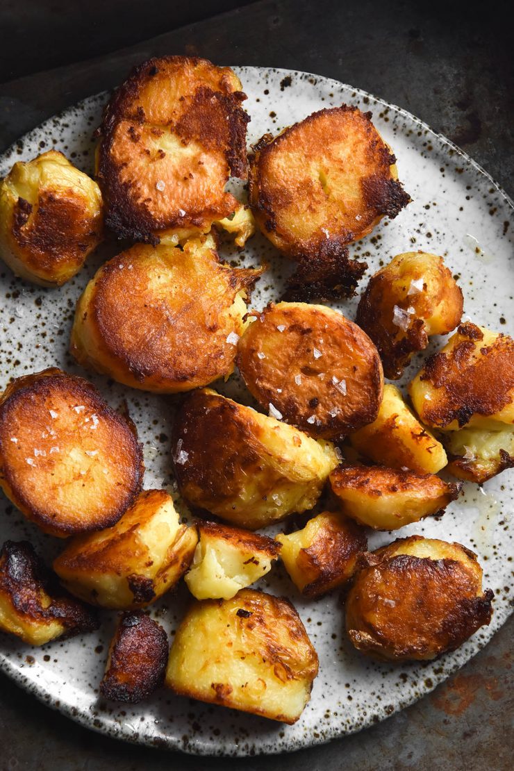 An aerial view of a plate of golden brown roast potatoes. The potatoes are sprinkled with sea salt flakes and sit atop a white speckled ceramic plate on a dark steel backdrop