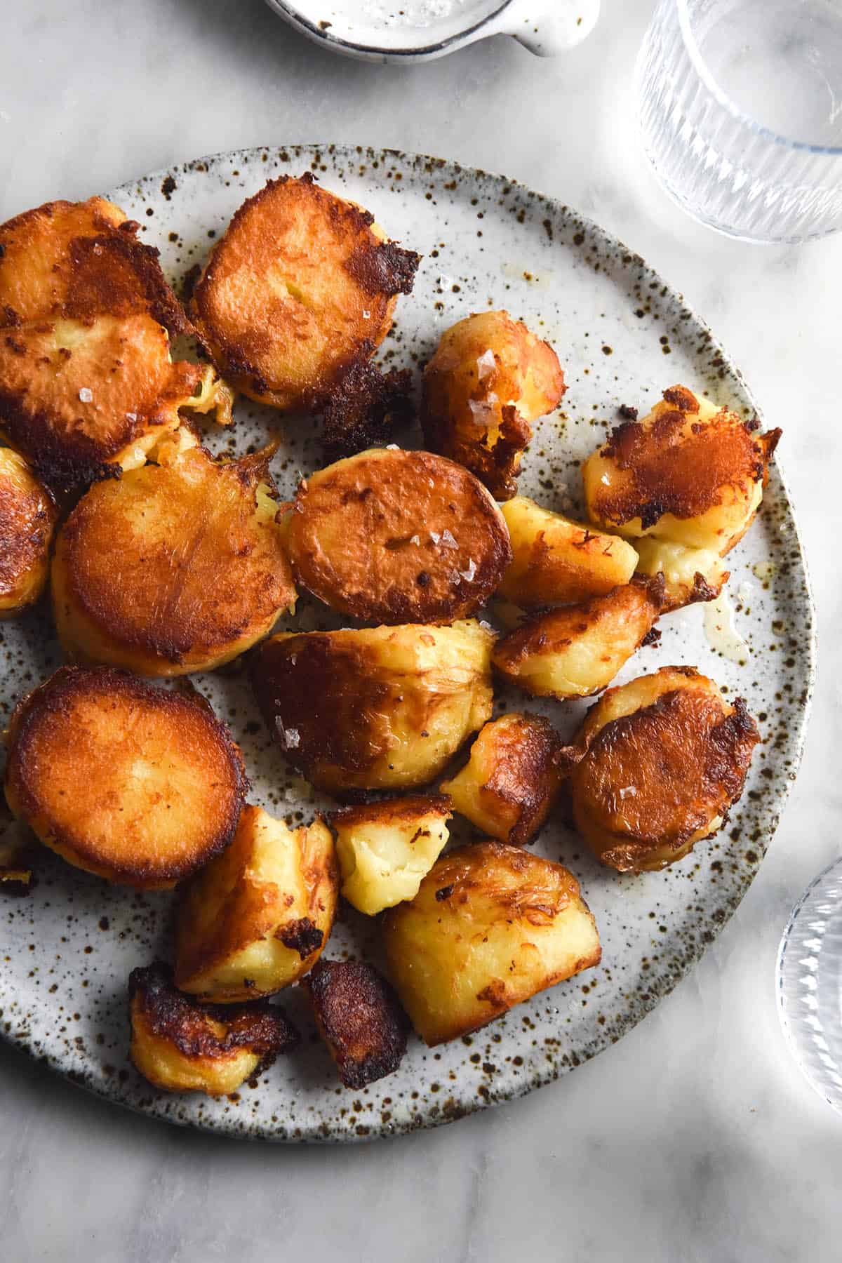 An aerial photo of a white speckled ceramic plate topped with crispy roast potatoes. The potatoes are golden brown, super crispy and topped with flaky sea salt. The scene is set on a white marble table with water glasses and salt bowls surrounding the plate of potatoes.