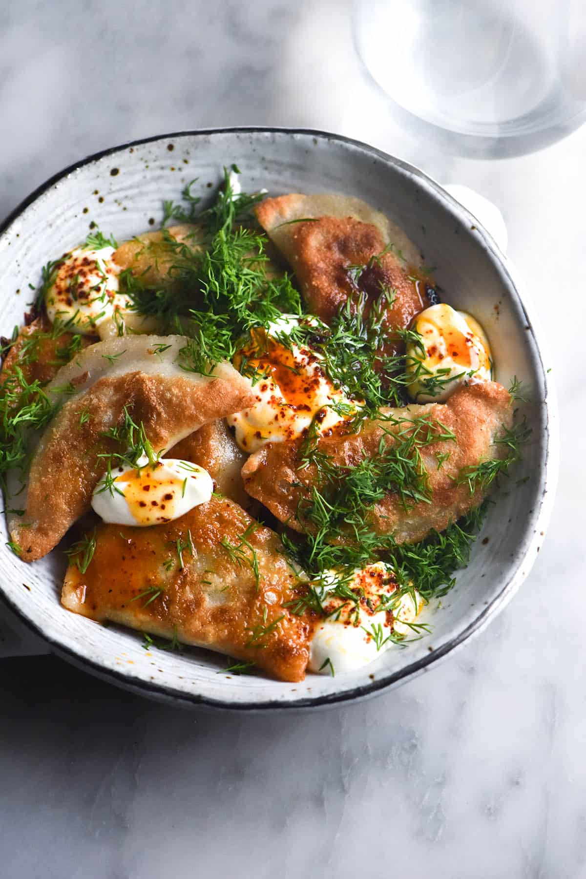 An aerial image of a white speckled ceramic bowl of gluten free Varenyky topped with sour cream, chilli oil and fresh dill. The bowl sits atop a white marble table. 