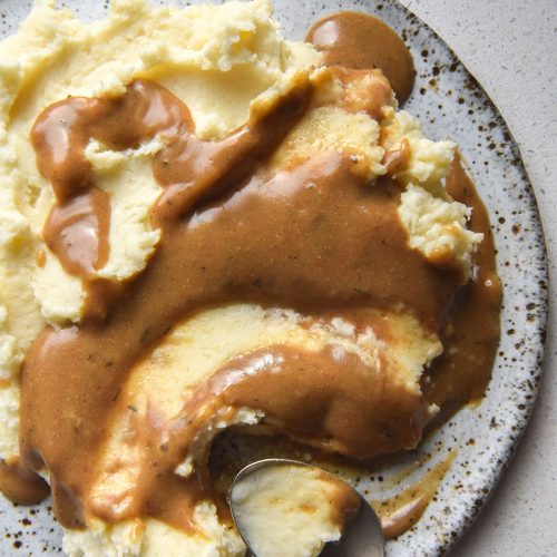 An aerial view of a plate of mashed potatoes smothered in FODMAP friendly vegan gravy. The dish sits atop a white ceramic plate on a white benchtop