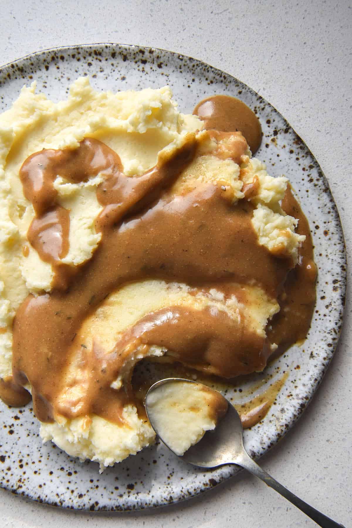 An aerial view of a plate of mashed potatoes smothered in FODMAP friendly vegan gravy. The dish sits atop a white ceramic plate on a white benchtop