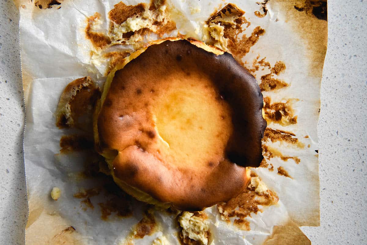 An aerial view of a mini Basque cheesecake on a white bench top. The top of the cheesecake is deeply caramelised and the browned baking paper has been peeled away from the cheesecake, revealing browned bits. 