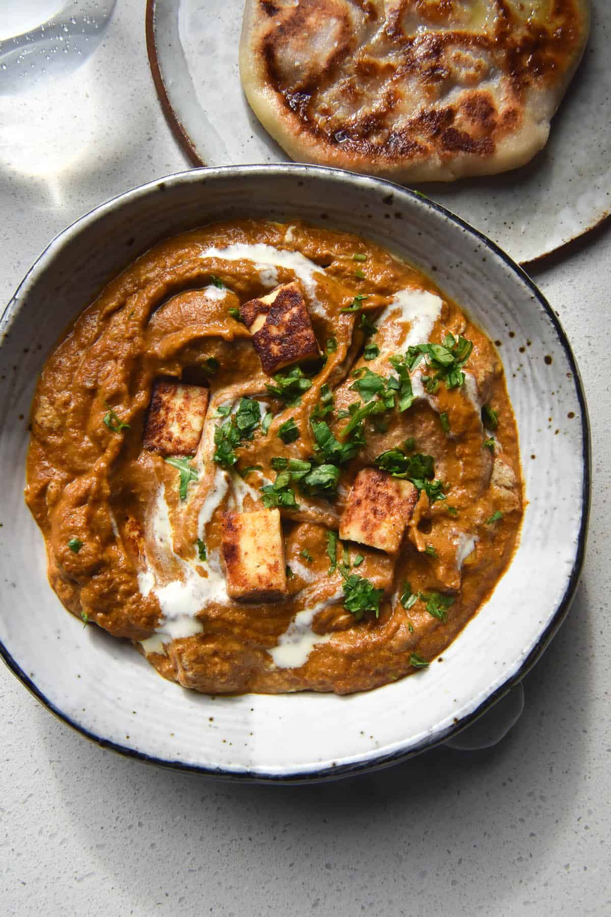 An aerial close up photo of a white speckled ceramic bowl filled with FODMAP friendly paneer curry. The curry is topped with a swirl of cream and chopped coriander, and some cubes of paneer peek out of the gravy. The bowl sits atop a white benchtop and a gluten free naan sits to the top right of the image. The image was been shot in bright sunlight, so the glass of water in the top left of the images casts light down onto the benchtop