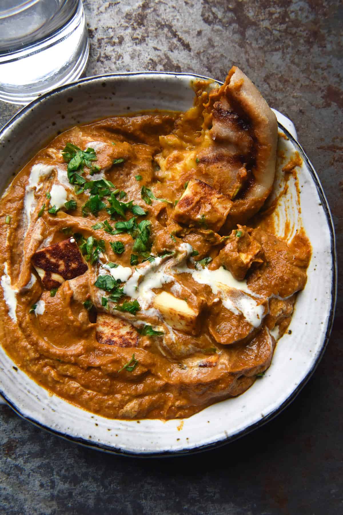 An aerial view of a white speckled ceramic bowl filled with FODMAP friendly paneer curry. A gluten free cheesy naan is being dipped into the top right corner of the curry, which is topped with a swirl of cream and chopped coriander. The bowl sits atop a blue steel backdrop and a glass of water sits to the top left of the image. 