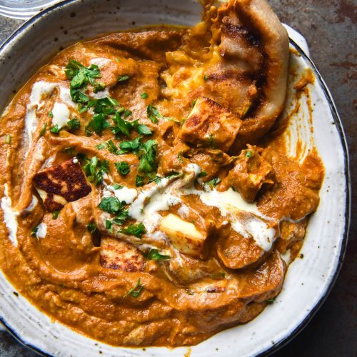 An aerial view of a bowl of FODMAP friendly paneer curry topped with a swirl of cream and chopped herbs. Paneer peeks out from the curry gravy and a cheesy naan dips into the top right corner of the curry. The bowl sits atop a grey steel backdrop and a glass of water sits to the top left of the image