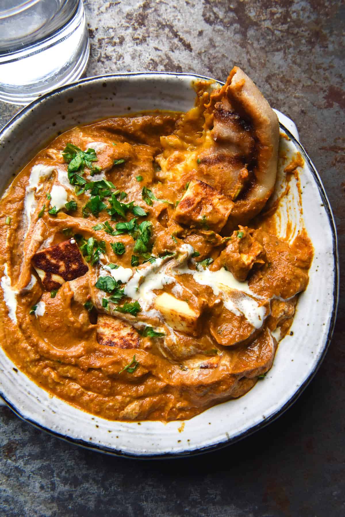 An aerial view of a bowl of FODMAP friendly paneer curry topped with a swirl of cream and chopped herbs. Paneer peeks out from the curry gravy and a cheesy naan dips into the top right corner of the curry. The bowl sits atop a grey steel backdrop and a glass of water sits to the top left of the image