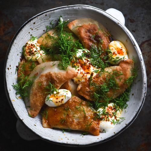 An aerial view of a white ceramic bowl filled with gluten free, FODMAP friendly Varenyky. They have been pan fried, with golden brown crispy surfaces. They are topped with sour cream, dill and chilli oil. The bowl sits atop a dark grey metal backdrop.