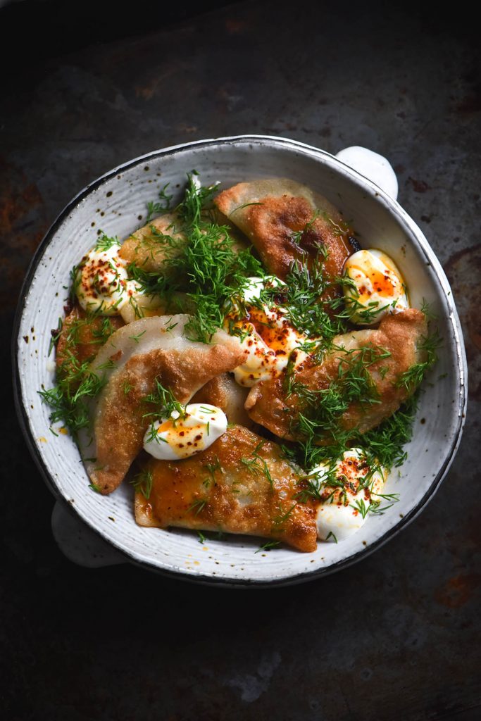 An aerial view of a white ceramic bowl filled with gluten free, FODMAP friendly Varenyky. They have been pan fried, with golden brown crispy surfaces. They are topped with sour cream, dill and chilli oil. The bowl sits atop a dark grey metal backdrop.