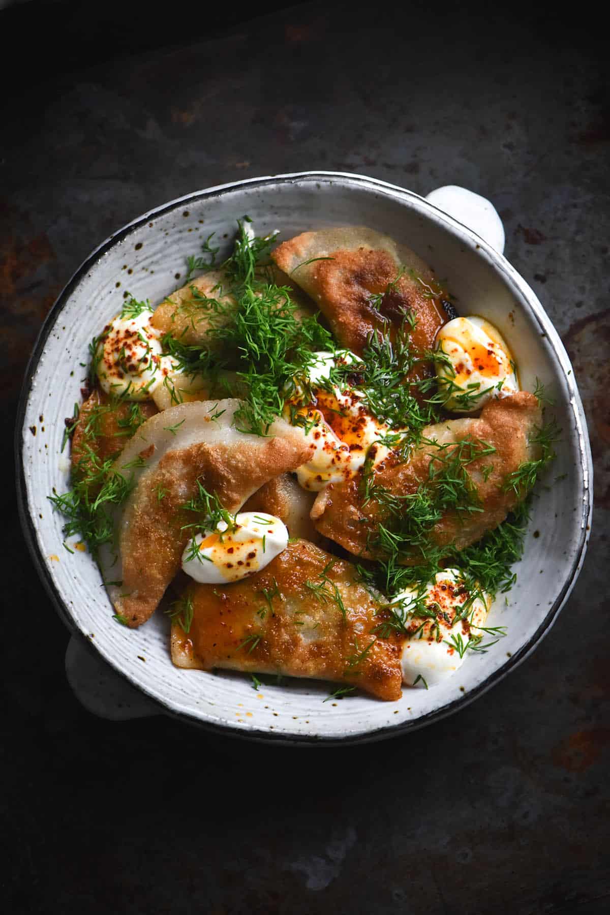 An aerial view of a white ceramic bowl filled with gluten free Varenkyky with a potato and cheese filling. The dumplings have been pan fried creating crispy golden edges. They are topped with sour cream, dill and chilli oil. The bowl sits atop a dark grey mottled background.