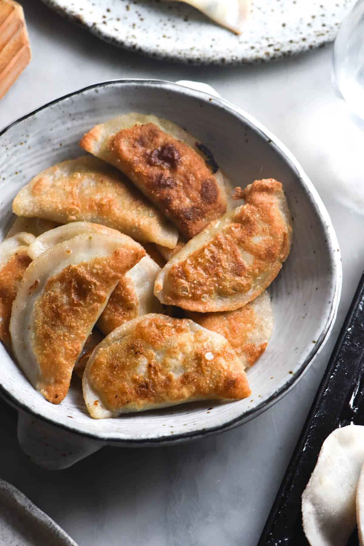 An aerial view of a bowl filled with crispy fried Varenkyky that are gluten free and filled with a FODMAP friendly leek, potato and cheese filling. The Vareniki are crispy and golden brown. The bowl sits on a white marble table surrounded by trays of uncooked vareniki.