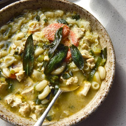 An aerial view of a beige speckled ceramic bowl filled with vegan chicken noodle soup. The soup is topped with crispy sage leaves, parmesan and truffle oil. It sits atop a white bench top in sunlight, and a glass of water streams light onto the top right of the image