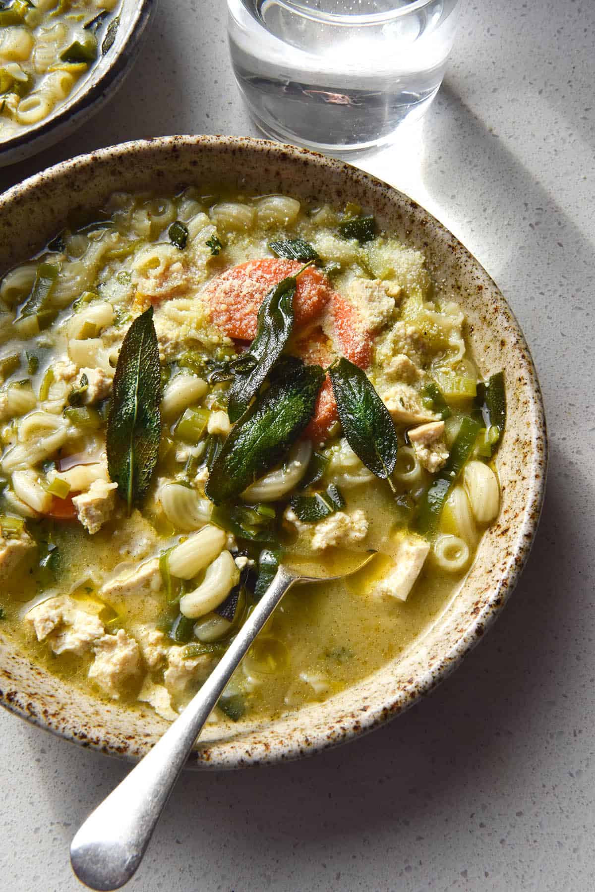 An aerial view of a beige speckled ceramic bowl filled with vegan chicken noodle soup. The soup is topped with crispy sage leaves, parmesan and truffle oil. It sits atop a white bench top in sunlight, and a glass of water streams light onto the top right of the image