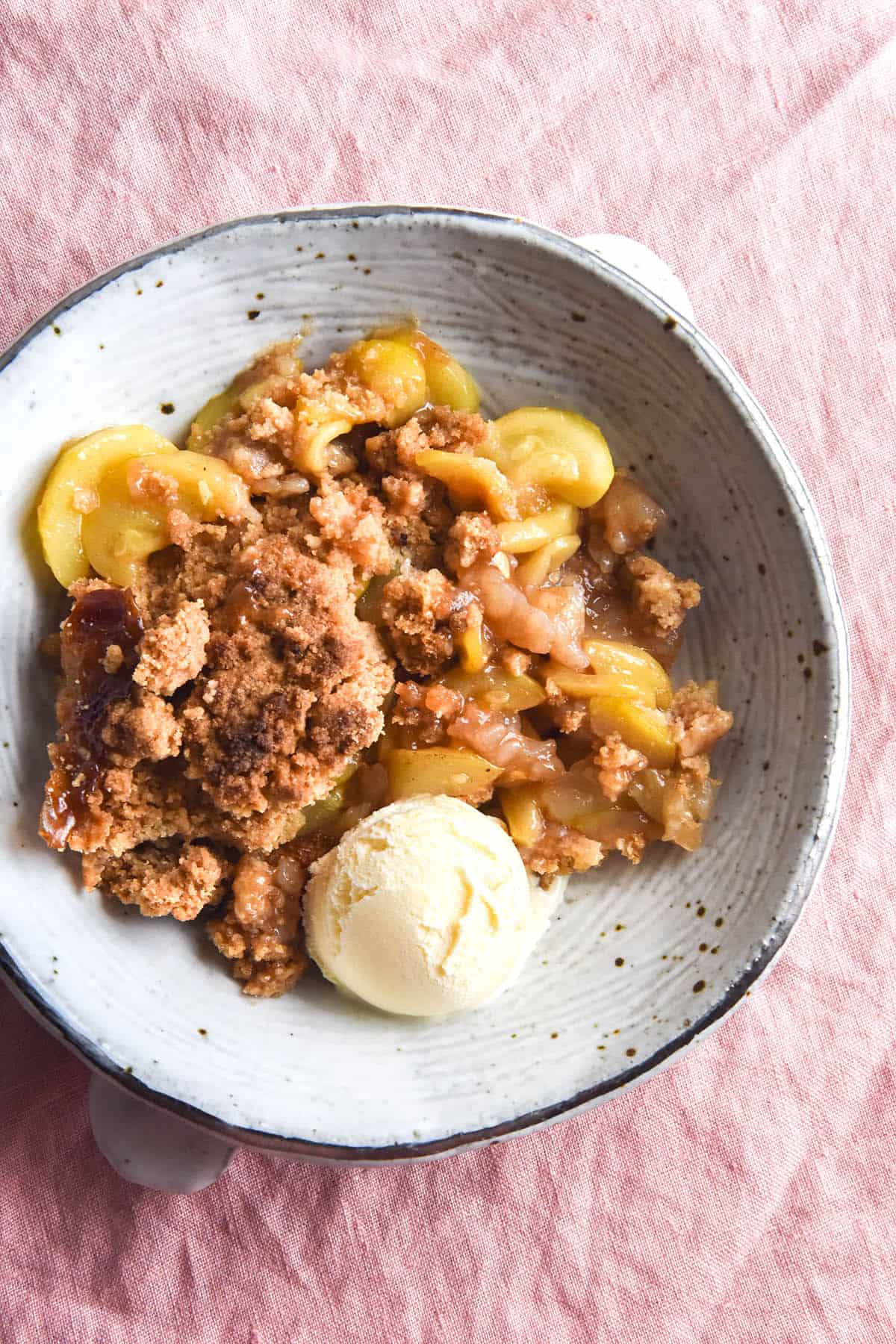 An aerial view of a white ceramic bowl filled with gluten-free zucchini 'apple' crumble and a scoop of vanilla ice cream. The bowl sits slightly off centre atop a light pink linen tea towel. 