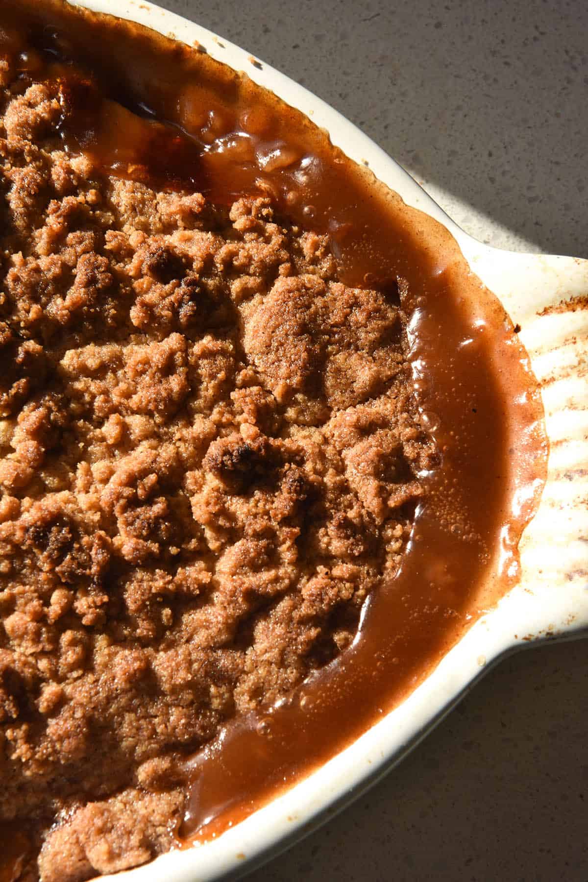 A close up aerial view of a gluten-free zucchini 'apple' crumble fresh from the oven. The crumble is deeply golden and the caramel from the filling has oozed up around the edges of the crumble. The crumble sits in a white baking dish atop a white bench top in bright sunlight. 