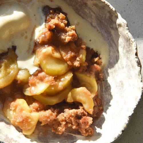 An aerial view of a bowl of gluten-free zucchini 'apple' crumble with a scoop of vanilla ice cream. It sits atop a white bench top in bright sunlight