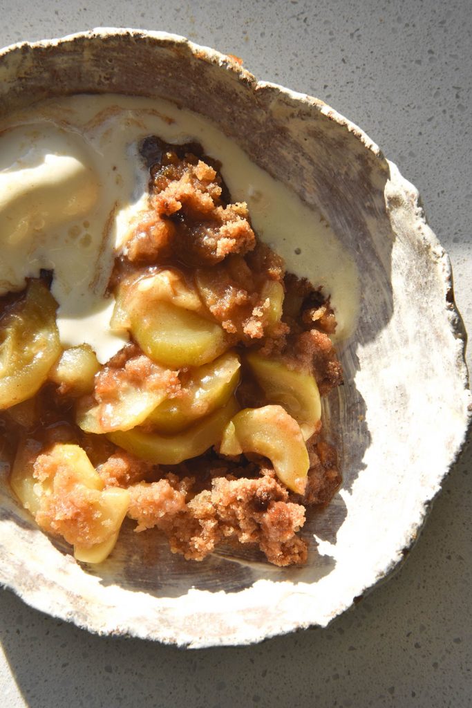 An aerial view of a bowl of gluten-free zucchini 'apple' crumble with a scoop of vanilla ice cream. It sits atop a white bench top in bright sunlight