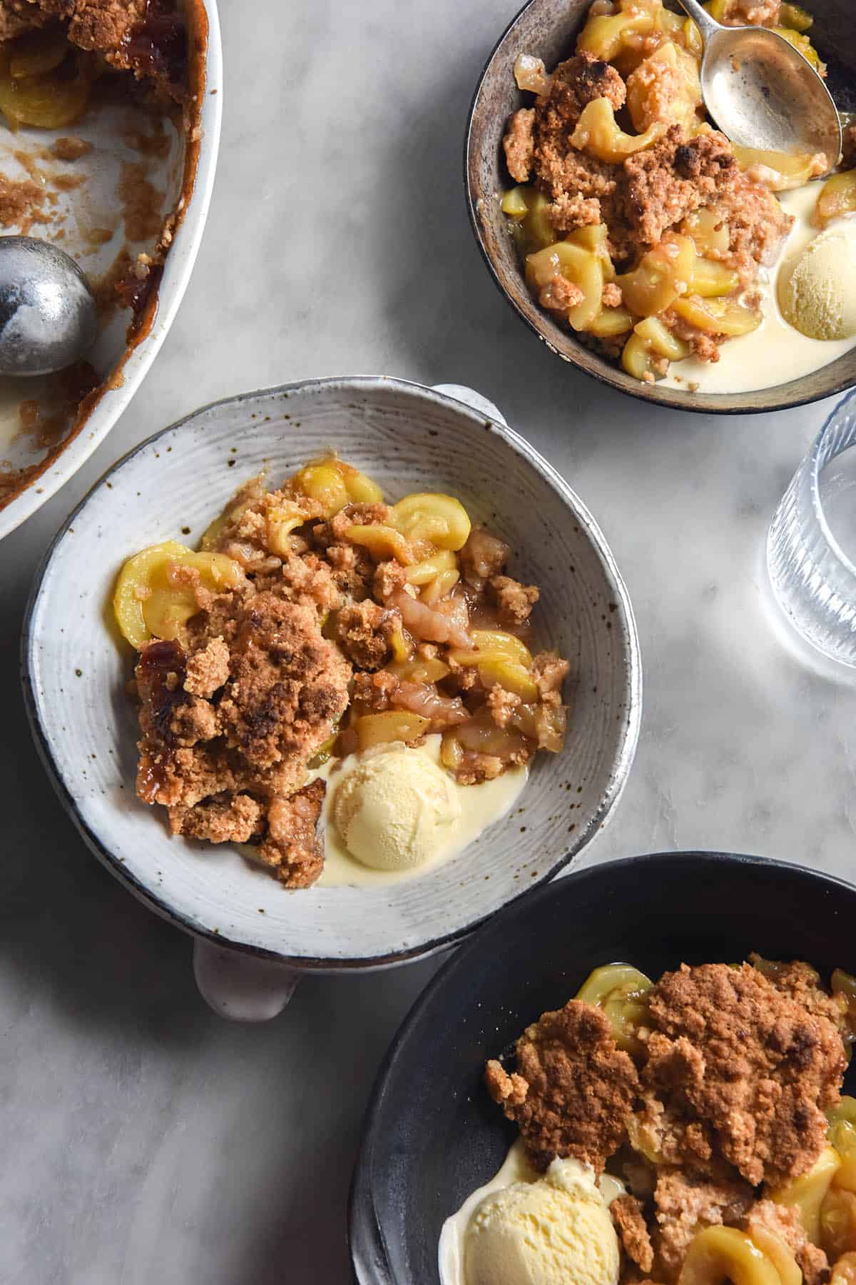 An aerial view of three ceramic plates topped with gluten-free zucchini 'apple' crumble atop a white marble table. The bowls are casually arranged on the table along with water glasses that the sun is streaming through