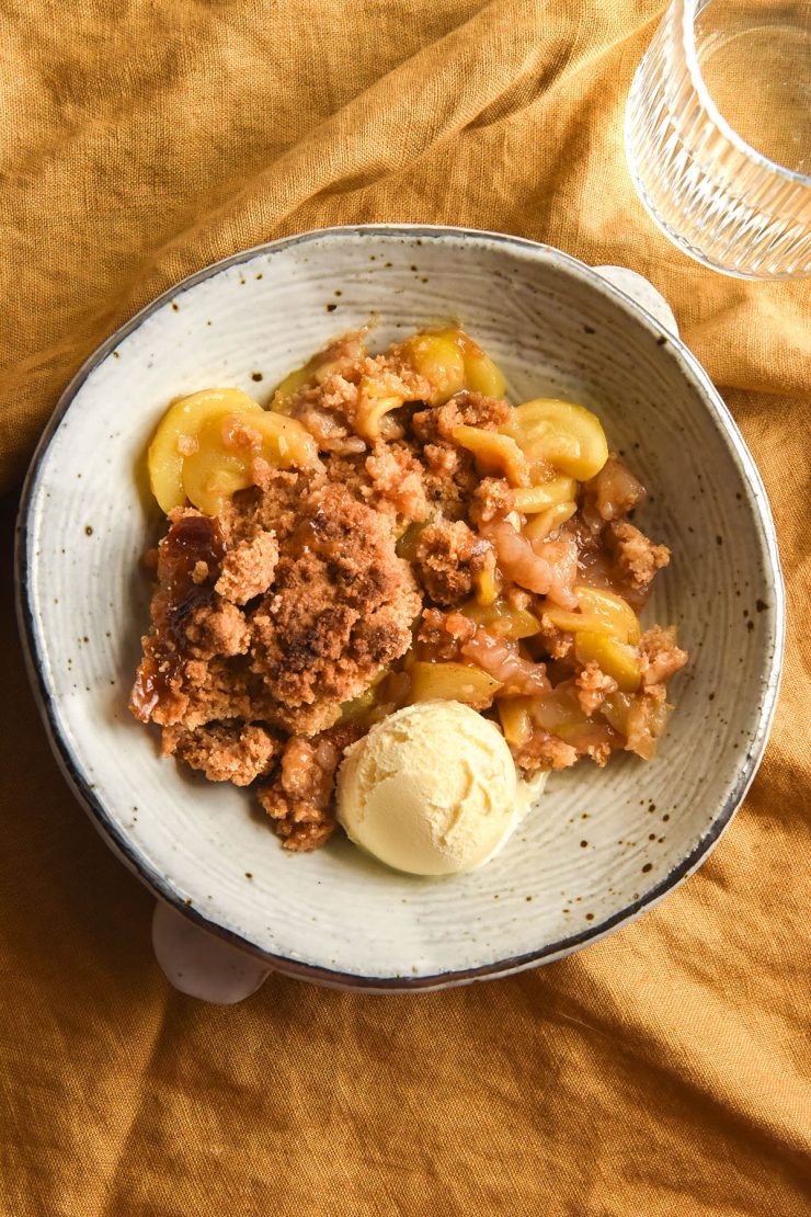 An aerial view of a white ceramic bowl filled with gluten free zucchini 'apple' crumble with a scoop of vanilla ice cream. The bowl sits atop a mustard coloured linen tablecloth and a glass of water sits to the top right of the image.