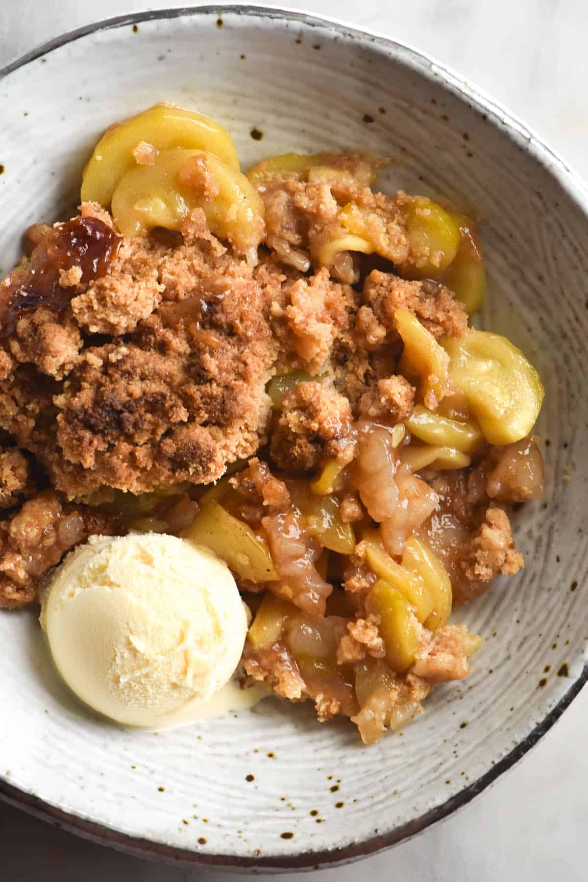 A close up aerial image of a gluten free zucchini 'apple' crumble with a scoop of vanilla ice cream in a white ceramic bowl. 