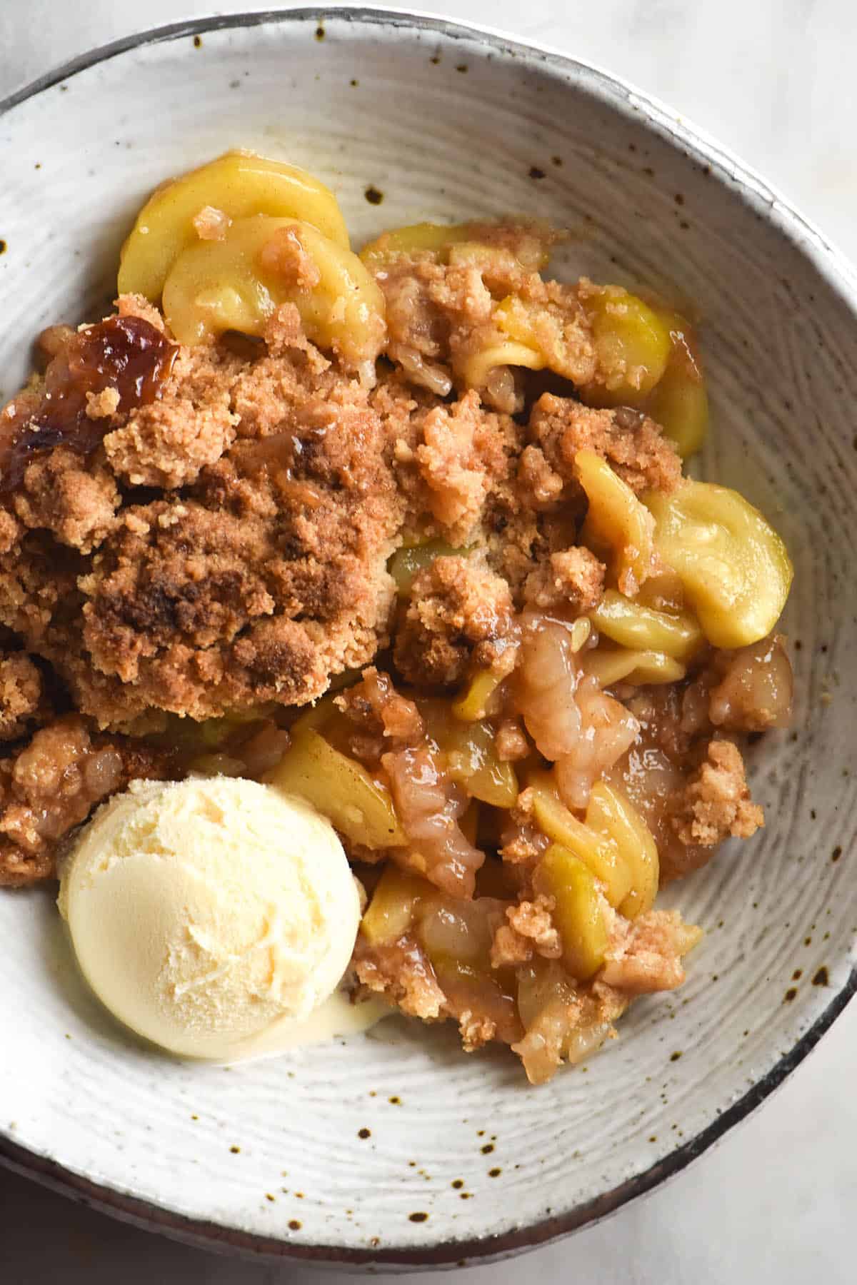 An aerial photo of a white speckled ceramic bowl filled with a zucchini 'apple' crumble that is gluten free. A scoop of vanilla ice cream sits to the bottom left of the bowl, which sits atop a white marble table. 