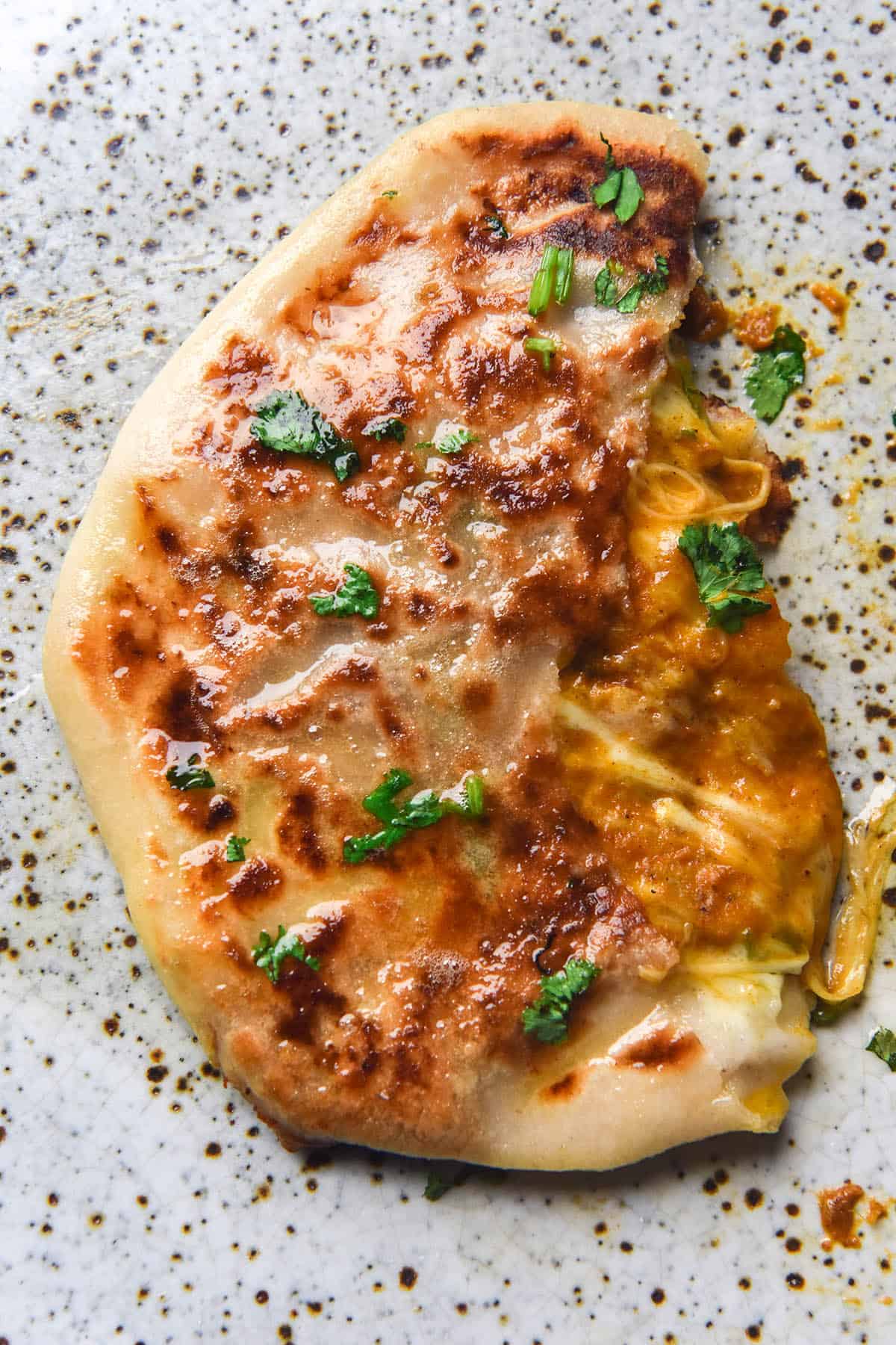 A gluten free cheesy naan that has been filled with melty cheese and a spoonful of FODMAP friendly curry. The flatbread sits atop a white speckled ceramic plate and is torn in half, exposing the melty cheese and curry innards. The flatbread has been topped with garlic infused ghee and chopped coriander.