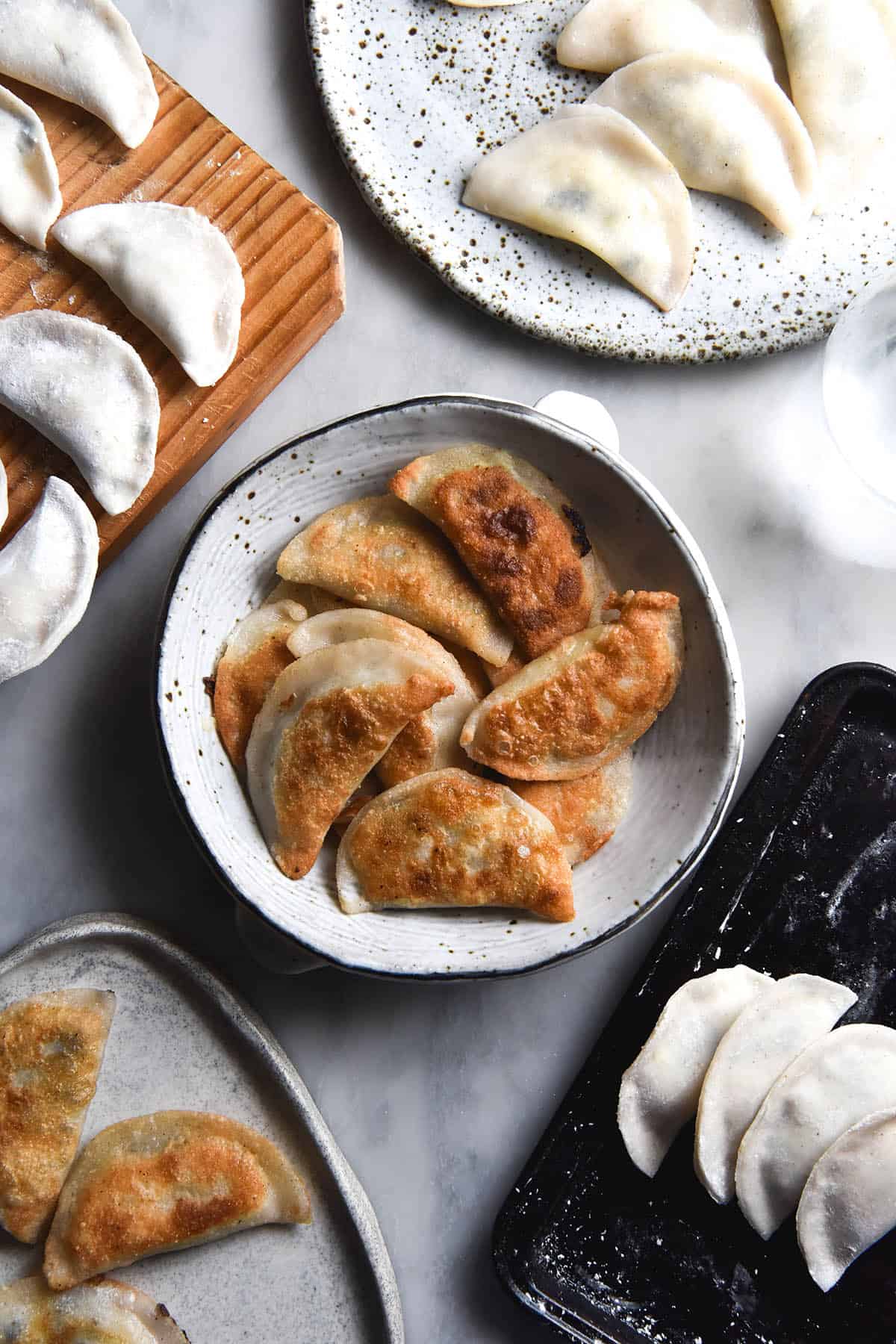 An aerial view of a white speckled ceramic bowl filled with golden brown fried varenyky that are gluten free. The bowl sits atop a white marble table and is surrounded by trays of uncooked Vareniki and plates of cooked Vareniki.