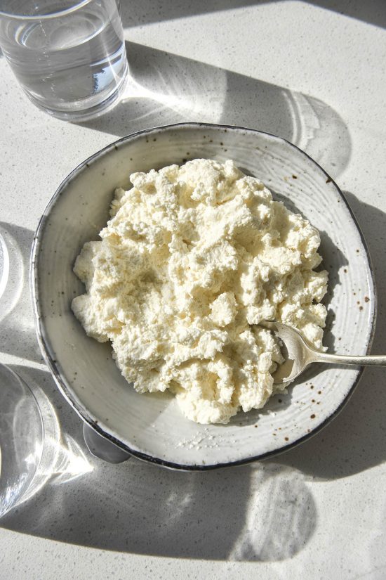 An aerial image of a white speckled ceramic bowl filled with lactose free cottage cheese sitting atop a white stone bench top. The bench is bathed in sunlight and three glasses of water sit to the left of the image, creating a light and shadow pattern across the image.
