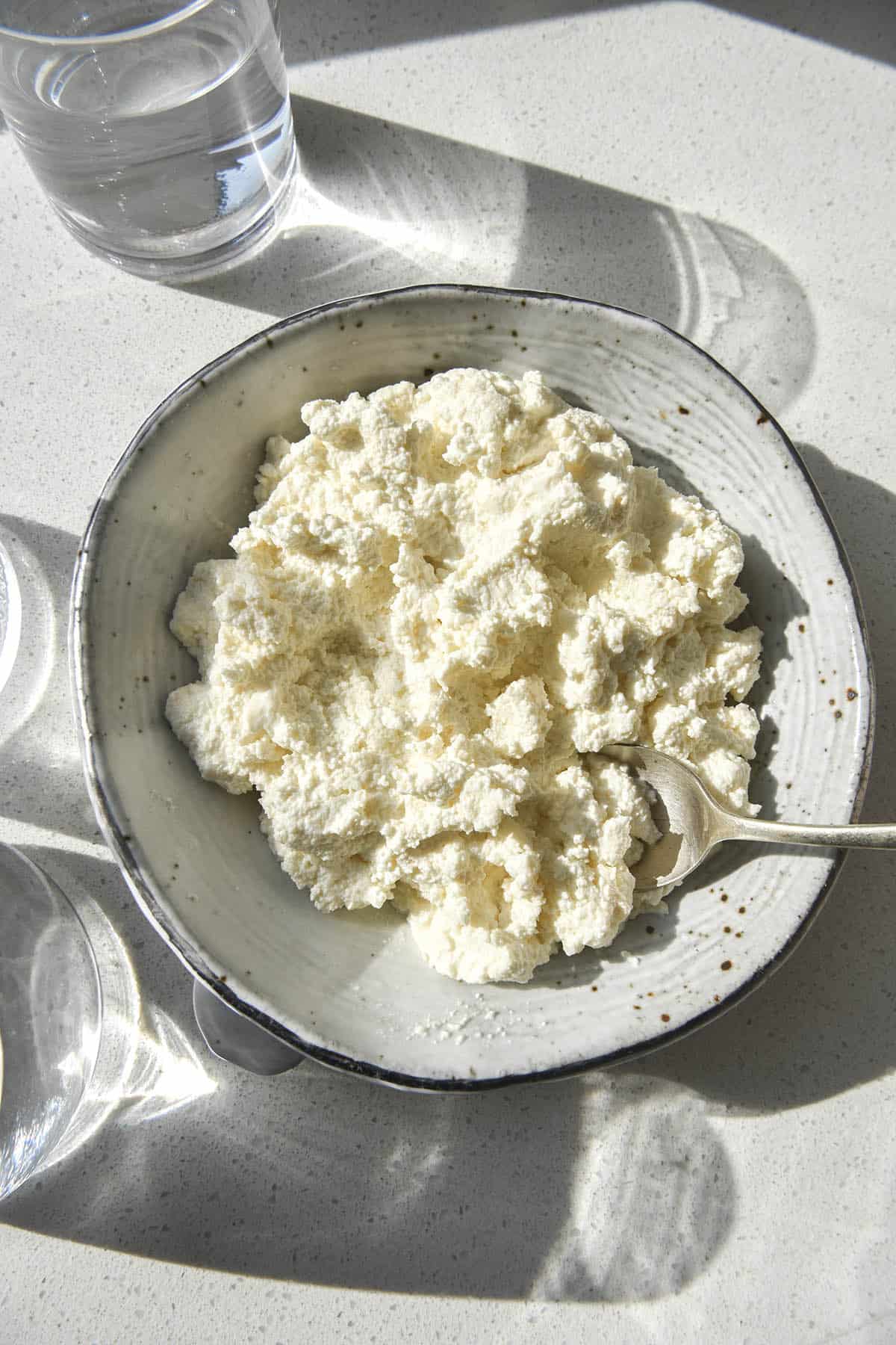 An aerial photo of a white ceramic speckled bowl filled with lactose free cottage cheese sitting atop a white stone bench top. Glasses of water sit to the left of the bowl and the sun streams through them, creating a light and shadow pattern across the image.