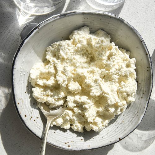 An aerial sunlit view of homemade lactose free cottage cheese. The cottage cheese is in a white speckled ceramic bowl surrounded by water glasses which create a light and shadow pattern over the image