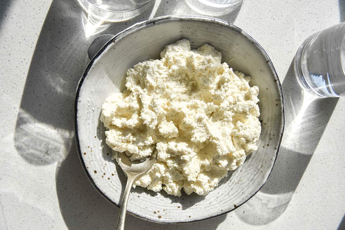 An aerial sunlit view of homemade lactose free cottage cheese. The cottage cheese is in a white speckled ceramic bowl surrounded by water glasses which create a light and shadow pattern over the image