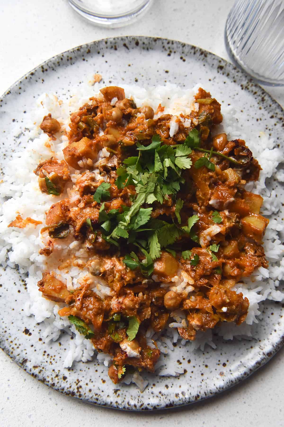 An aerial view of a white speckled ceramic plate topped with rice and FODMAP friendly vegetable tagine topped with coriander. The plate sits atop a white benchtop and is surrounded by water glasses.