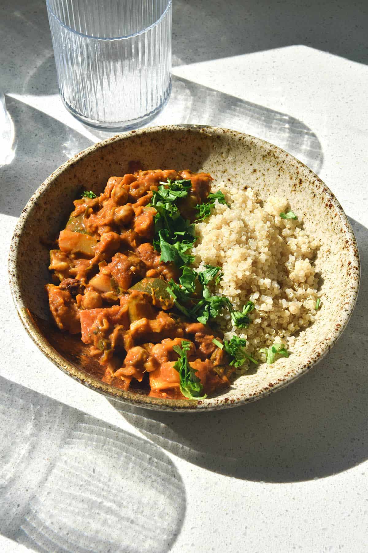 A brightly sunlit view of a speckled ceramic bowl of FODMAP friendly vegetable tagine served with quinoa and topped with coriander. The bowl sits on a white stone benchtop and is surrounded by glasses of water that create a shadow over the image
