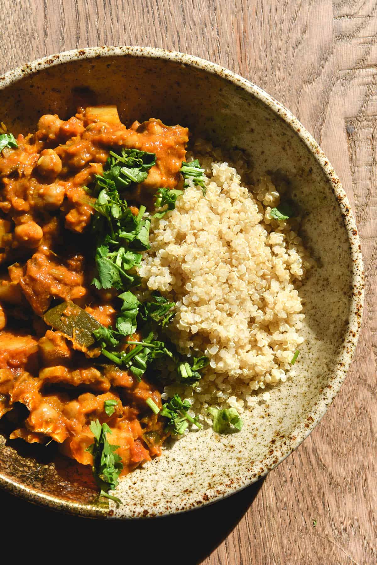 An aerial sunlit view of a speckled ceramic bowl filled with FODMAP friendly tagine and quinoa topped with coriander. The bowl sits on a wooden backdrop in the sun.