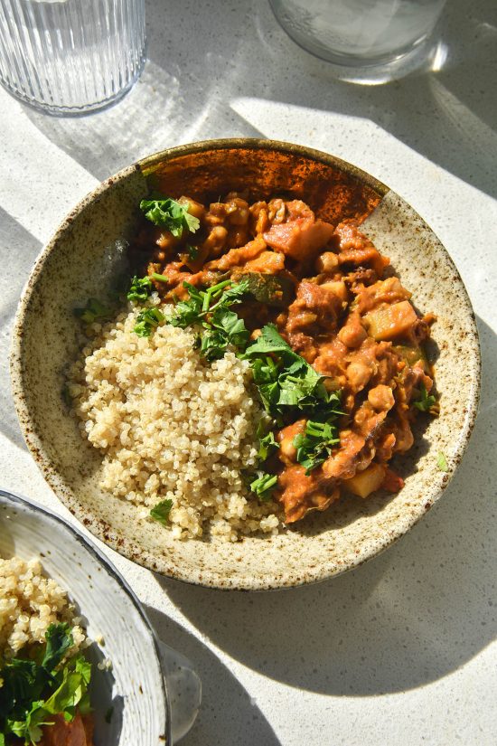 An aerial sunlit view of a speckled ceramic bowl filled with FODMAP friendly vegetable tagine and quinoa topped with coriander. The bowl sits on a white stone benchtop surrounded by glasses of water and another bowl of tagine.